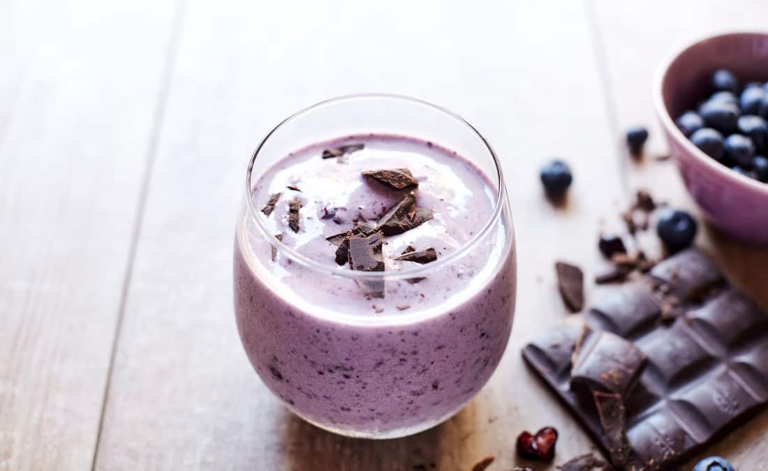 purple smoothie for beginners in a glass with blueberries, chocolate shavings, and a chocolate bar on a wooden table